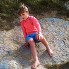 A child wearing a red UV Protective Rash Vest and blue starfish shorts sits on a rock, looking down and pointing with their right hand.